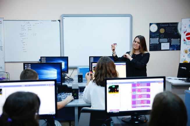 A teacher in front of a class holding up a micro:bit 